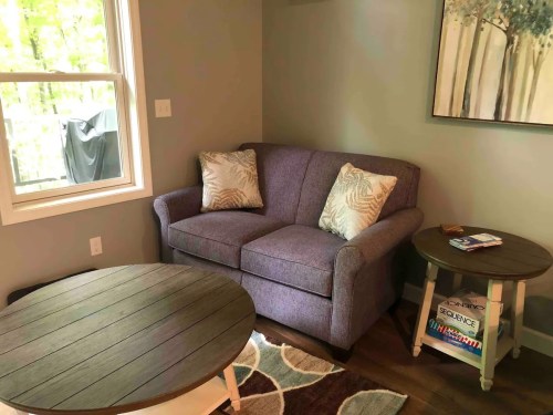Cozy corner with a purple loveseat, decorative pillows, a round coffee table, and a window with greenery outside.