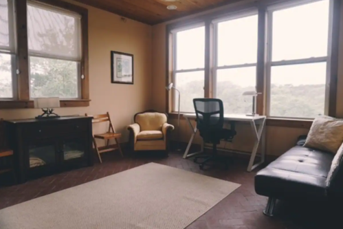 Cozy room with large windows, a desk, chair, armchair, and a rug on a wooden floor.