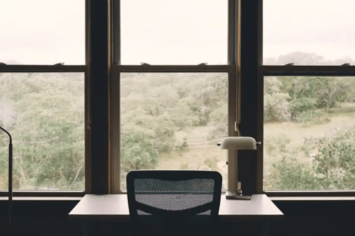A minimalist desk with a chair and lamp, facing large windows overlooking a green, tree-filled landscape.