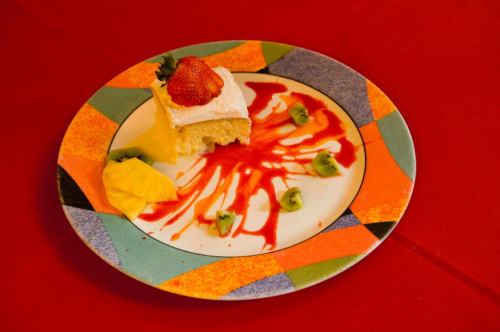 A slice of cake topped with cream and a strawberry, drizzled with red sauce, on a colorful plate with fruit pieces.