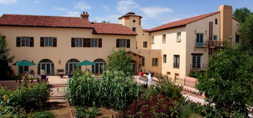 A sunny courtyard with lush gardens and a Spanish-style building featuring arched windows and a red tile roof.