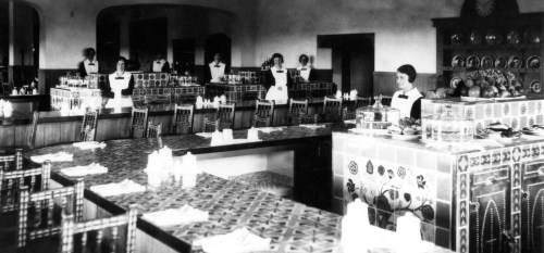 A historic dining room with several waitresses, tables set, and decorative tiles in a vintage style.
