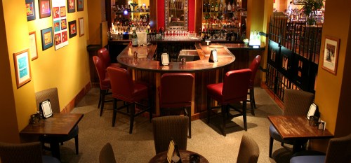 Cozy bar area with a curved counter, red chairs, and wooden tables, adorned with colorful artwork on the walls.