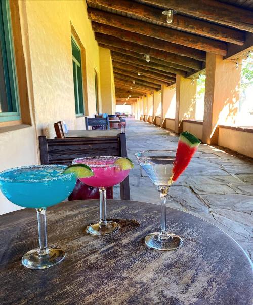 Three colorful cocktails on a wooden table, with a sunny corridor in the background.