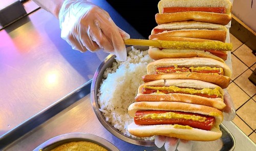A person in gloves holds a stack of hot dogs with mustard, next to a bowl of rice and a container of sauce.