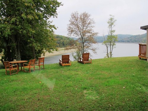 A serene lakeside view with wooden chairs and a table on a grassy area, surrounded by trees and distant hills.