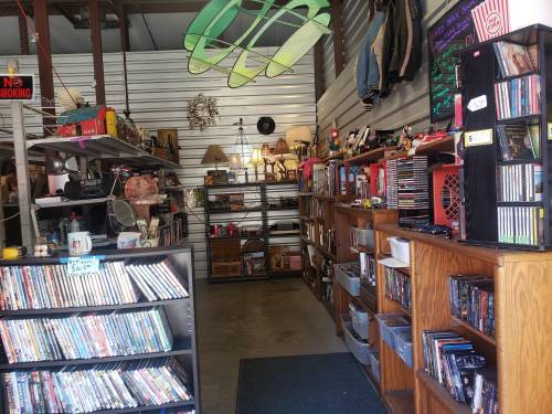 A cozy thrift store interior with shelves of DVDs, CDs, and various items, including lamps and clothing.