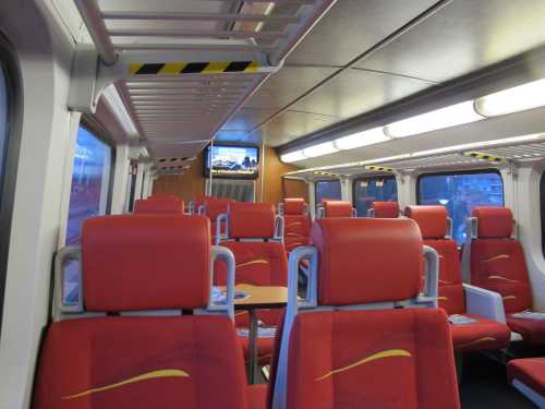 Interior of a train carriage with red seats, tables, and a screen displaying information. The space is empty.