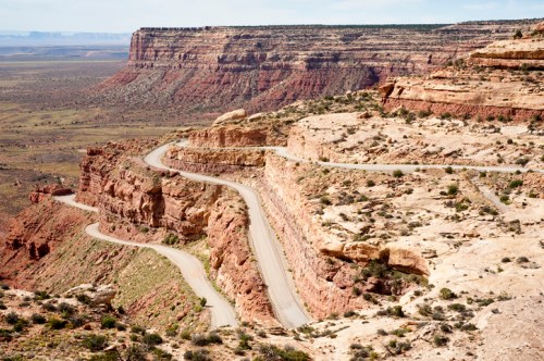 Winding road along a rocky cliff with expansive desert landscape in the background.