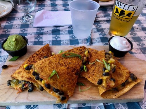 A wooden platter with four crispy quesadillas filled with cheese, black beans, and vegetables, served with guacamole and sour cream.