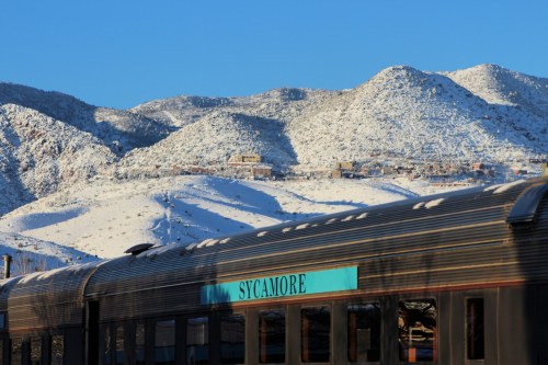 A train car labeled "Sycamore" in the foreground, with snow-covered mountains in the background under a clear blue sky.