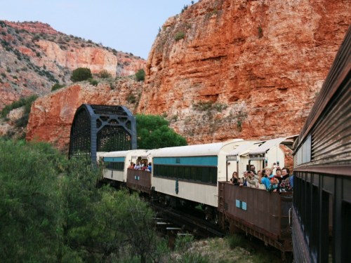 A vintage train crosses a bridge surrounded by red rock formations and greenery, with passengers enjoying the view.