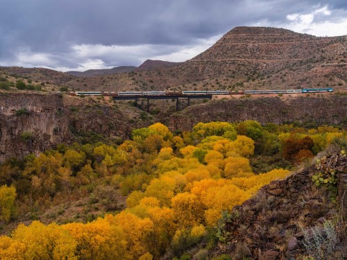 A train crosses a bridge over a canyon filled with vibrant autumn foliage under a cloudy sky.