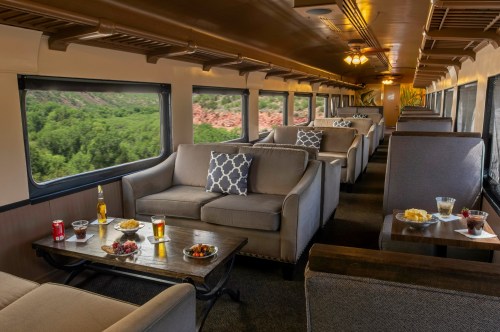 Interior of a train lounge with cozy seating, tables with snacks and drinks, and large windows showcasing greenery outside.