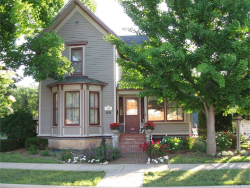 A charming gray Victorian house with a front porch, surrounded by green trees and colorful flower beds.