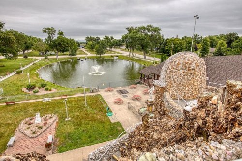 Aerial view of a park with a pond, fountain, and picnic area, surrounded by trees and a stone structure in the foreground.