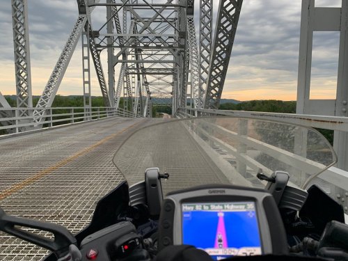 View from a motorcycle on a metal bridge, with a GPS display showing directions and a cloudy sunset in the background.