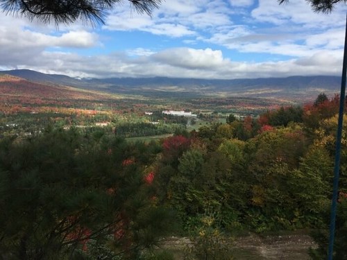 A scenic view of a valley with colorful autumn foliage, mountains in the background, and a partly cloudy sky.