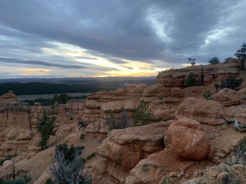 A rocky landscape at sunset, with layered cliffs and scattered trees under a cloudy sky.