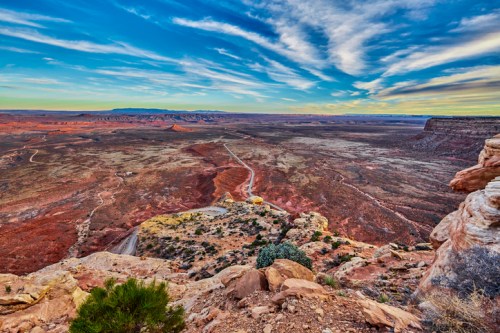A vast desert landscape with red rock formations and a winding road under a colorful sky.