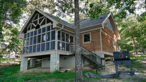 A wooden house with a screened porch, surrounded by trees, and a grill nearby on a grassy area.