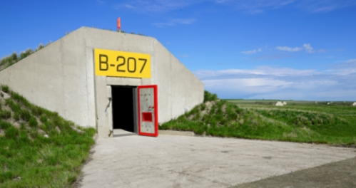 A concrete bunker with a yellow "B-207" sign and a red door, set against a grassy landscape and blue sky.