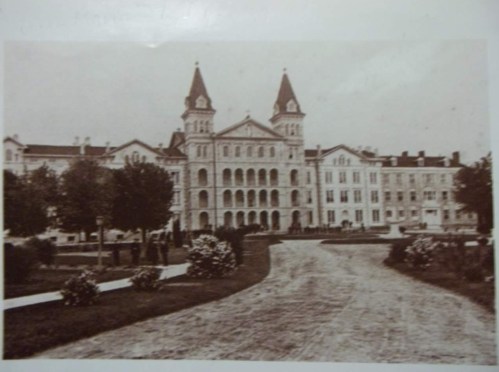 Historic black-and-white photo of a large building with towers, surrounded by trees and a pathway.