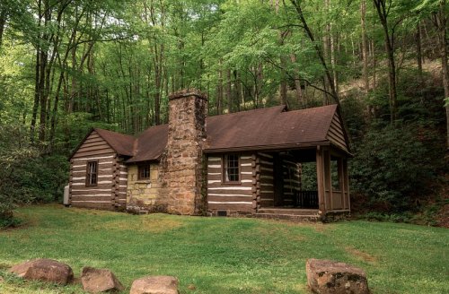 A rustic log cabin with a stone chimney, surrounded by lush green trees and grass.
