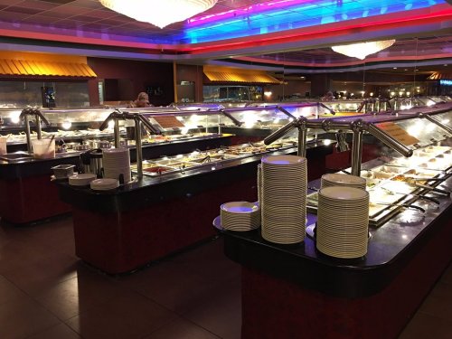 A brightly lit buffet with various food trays and stacked plates in a spacious dining area.