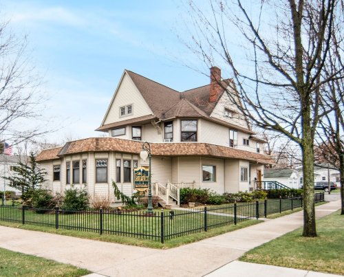 A large, charming house with a sloped roof, surrounded by greenery and a black fence, on a clear day.