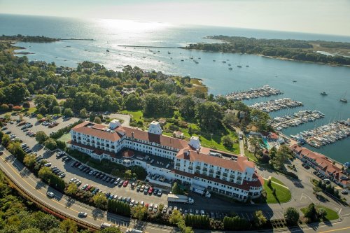 Aerial view of a coastal hotel near a marina, surrounded by trees and water, with boats docked in the harbor.