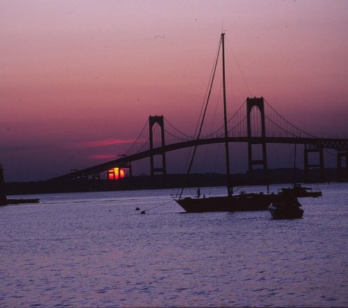 A sunset casts a warm glow behind a bridge, with a sailboat in the foreground on calm waters.