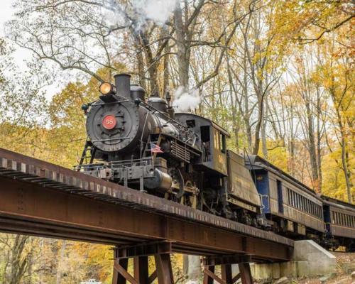 A vintage steam train crosses a bridge surrounded by colorful autumn trees. Smoke billows from the locomotive.