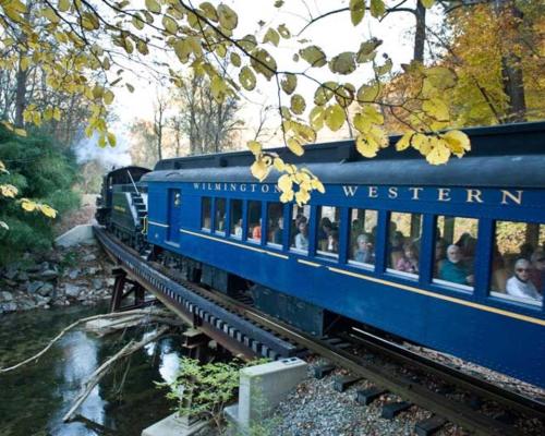 A blue train with passengers travels along a scenic track surrounded by autumn foliage and a small stream.