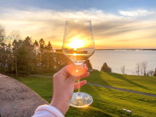 A hand holding a wine glass with the sun setting over a lake and trees in the background.