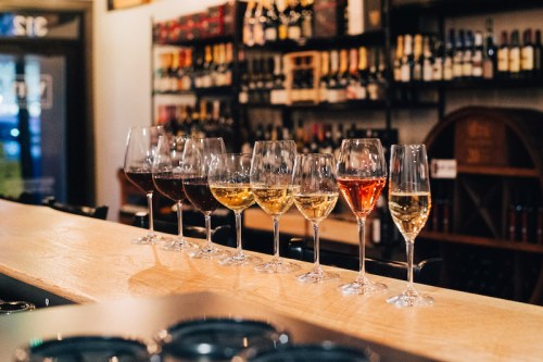A row of wine glasses filled with different types of wine on a bar, with a wine shelf in the background.