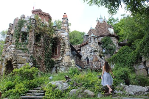 A woman in a flowing dress walks towards a whimsical stone castle surrounded by lush greenery and trees.