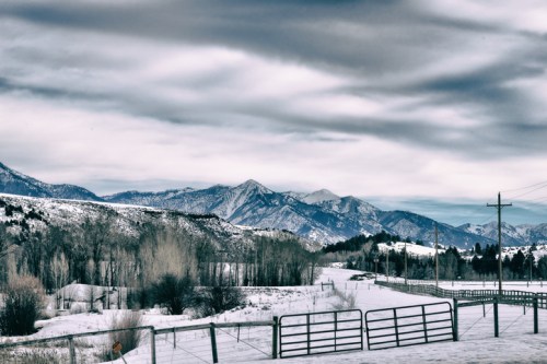 Snow-covered landscape with mountains in the background and a cloudy sky, featuring a fence and power lines.