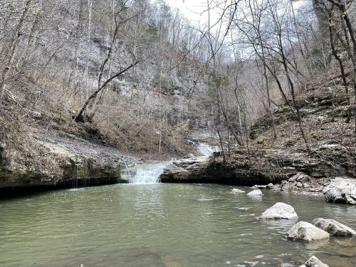 A serene landscape featuring a small waterfall cascading into a calm pool, surrounded by rocky terrain and bare trees.