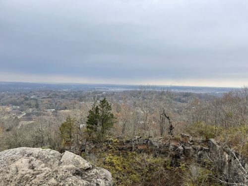 A rocky overlook with a view of a valley and distant city under a cloudy sky, surrounded by sparse trees.