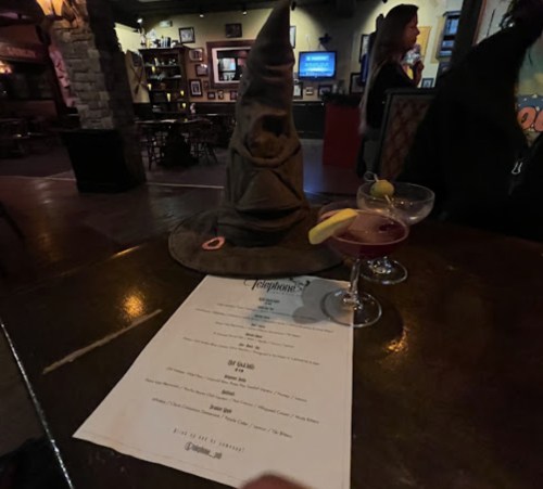 A wizard hat sits on a table next to two cocktails and a menu in a cozy bar setting.