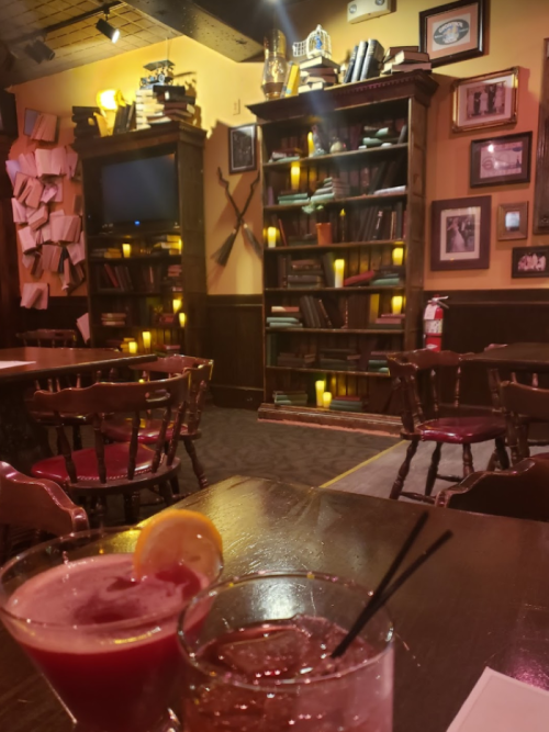 Cozy bar scene with drinks in foreground, bookshelves and warm lighting in the background, creating an inviting atmosphere.