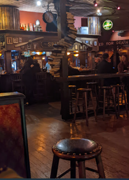 A cozy bar interior with wooden decor, a spiral of books, and patrons enjoying drinks at the counter.