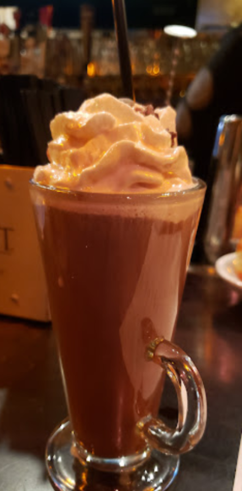 A glass of hot chocolate topped with whipped cream and a drizzle of caramel, served with a straw.