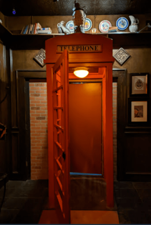 An orange telephone booth with an open door, set against a dark, decorated wall with shelves and framed pictures.