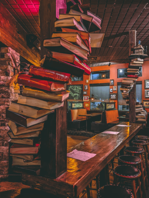 A cozy bar interior with books stacked creatively on wooden pillars and warm lighting, creating an inviting atmosphere.