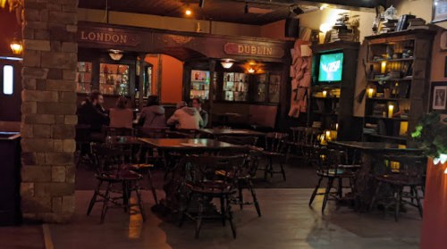 Cozy pub interior with wooden tables, chairs, and shelves, featuring a warm ambiance and a TV in the background.