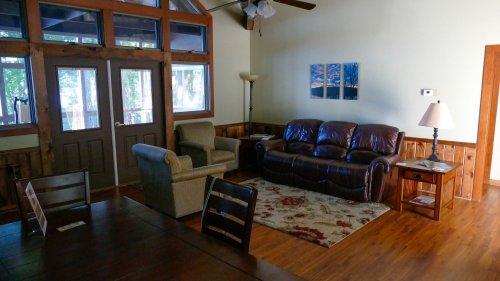 Cozy living room with a leather sofa, armchair, wooden dining table, and large windows letting in natural light.