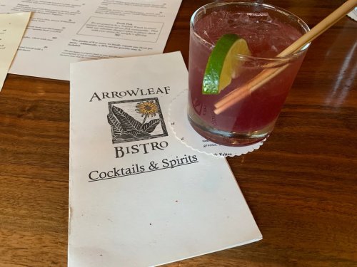 A cocktail with a lime wedge sits next to a menu for Arrowleaf Bistro, featuring cocktails and spirits.