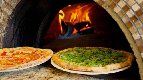 Two pizzas, one with tomato and cheese and the other topped with arugula, baking in a wood-fired oven.